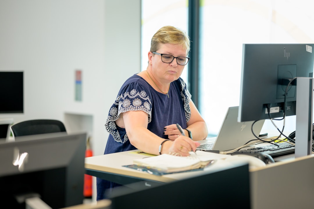 A woman working at a standing desk. There is a laptop in front of her and shes writing down something on a notepad.